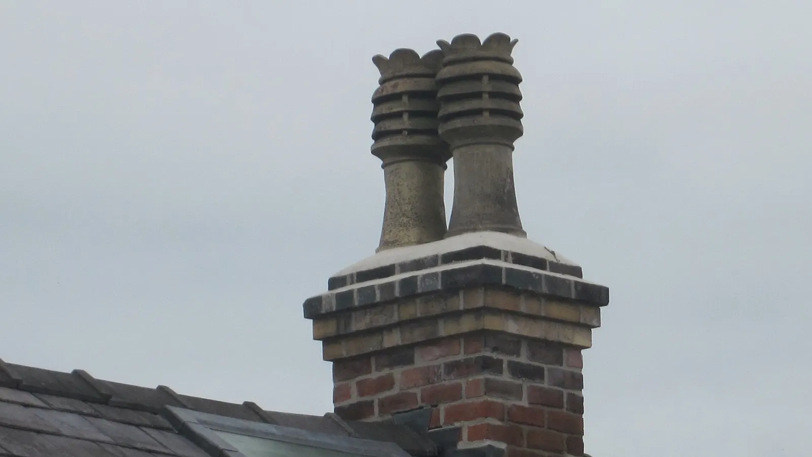 Brick chimney stack with two tall chimney pots rising above a slate roof against a pale sky