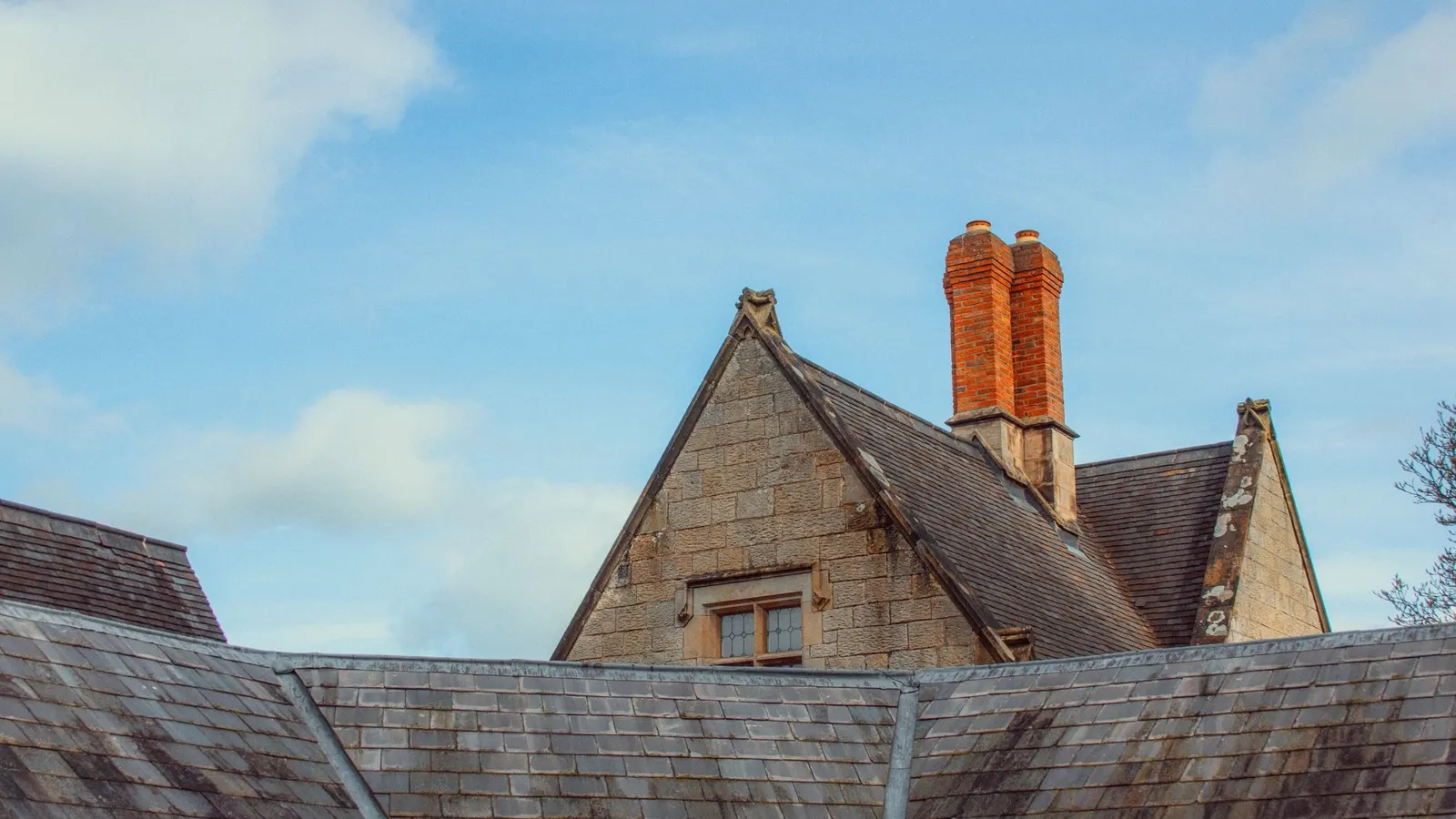 Stone house with steep slate roof and tall red brick chimney stack in bright daylight