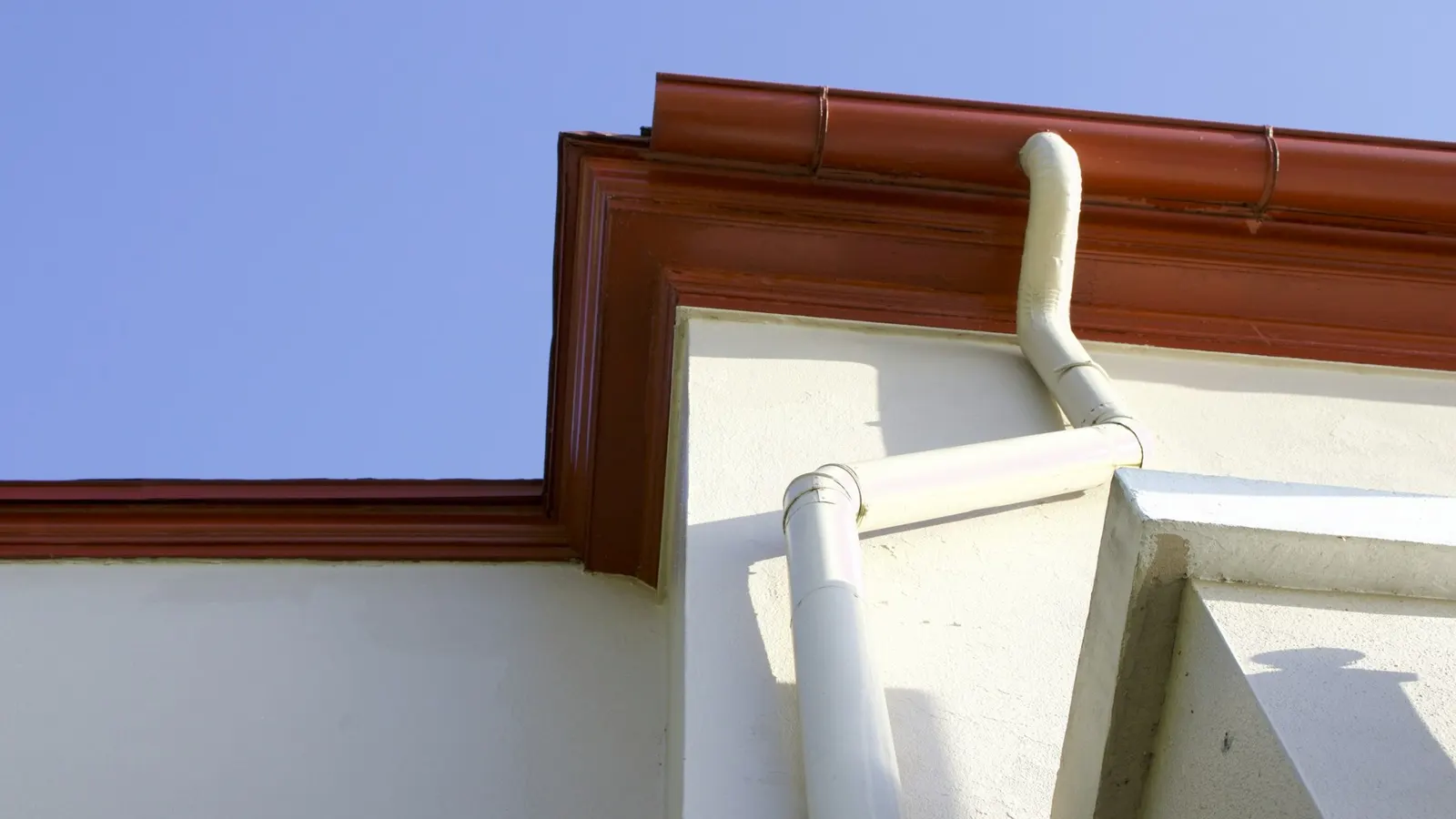 Brown gutter and fascia with white downpipe on the eaves of a house under blue sky