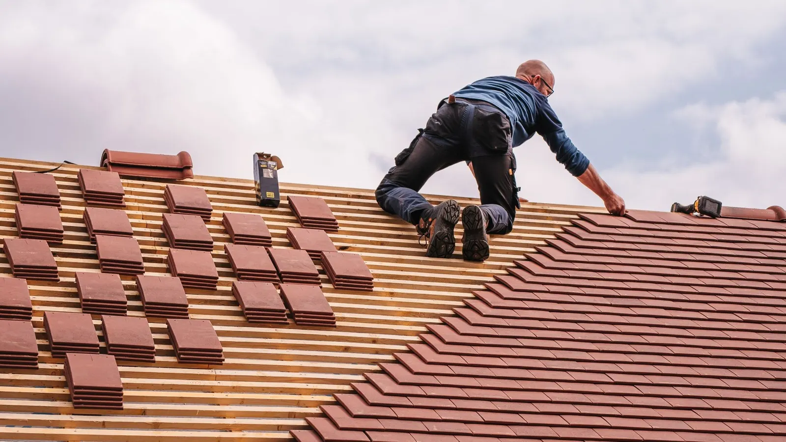 Roofer fitting new plain clay tiles on a pitched roof, with tile stacks ready on the battens