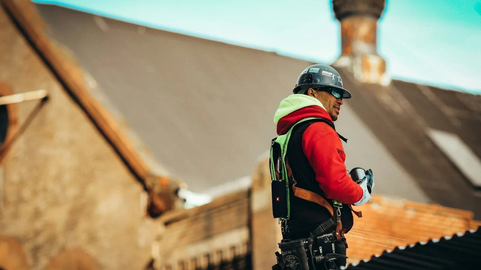 Roofer in hard hat and safety harness inspecting a pitched tiled roof beside a brick chimney