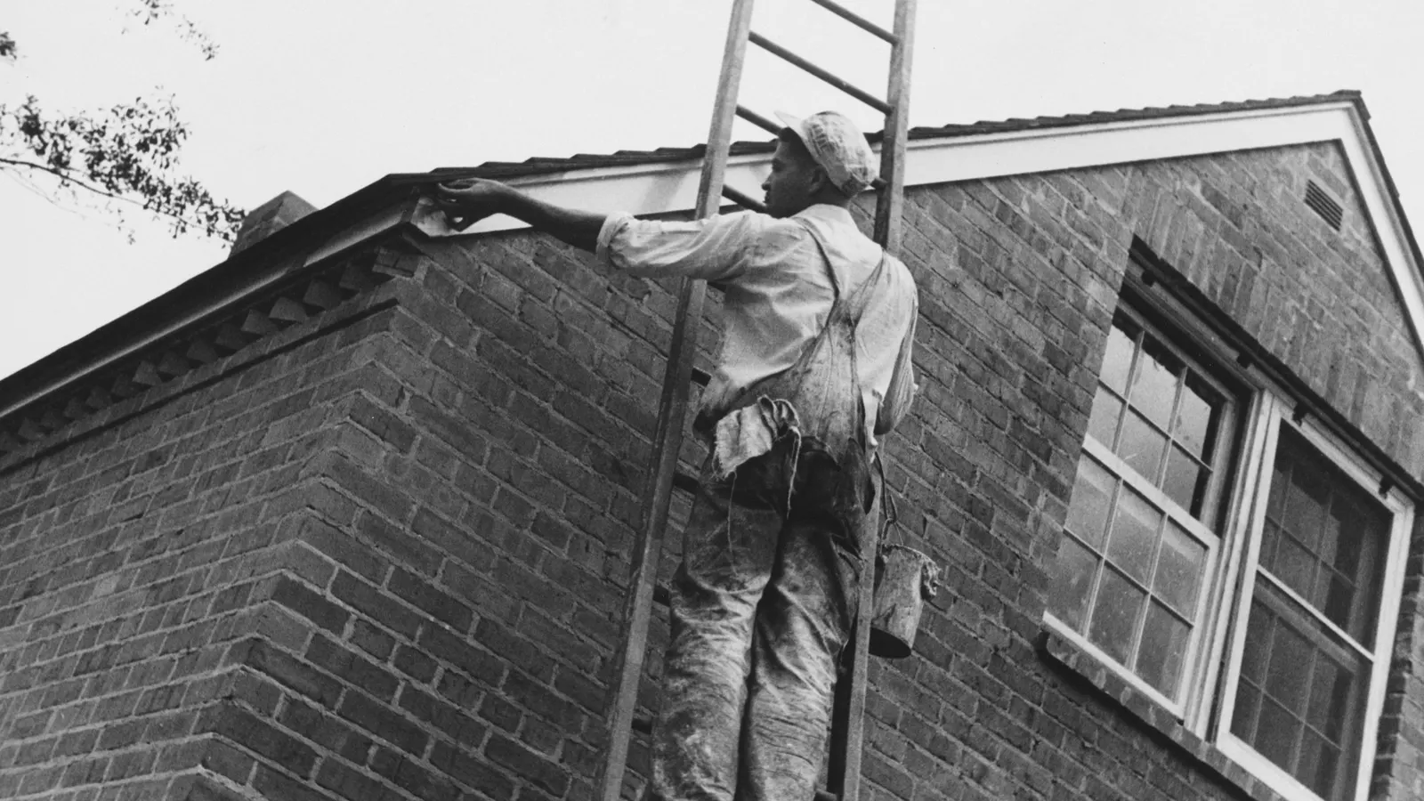 Tradesman on a ladder against a brick house, checking the roof edge and guttering